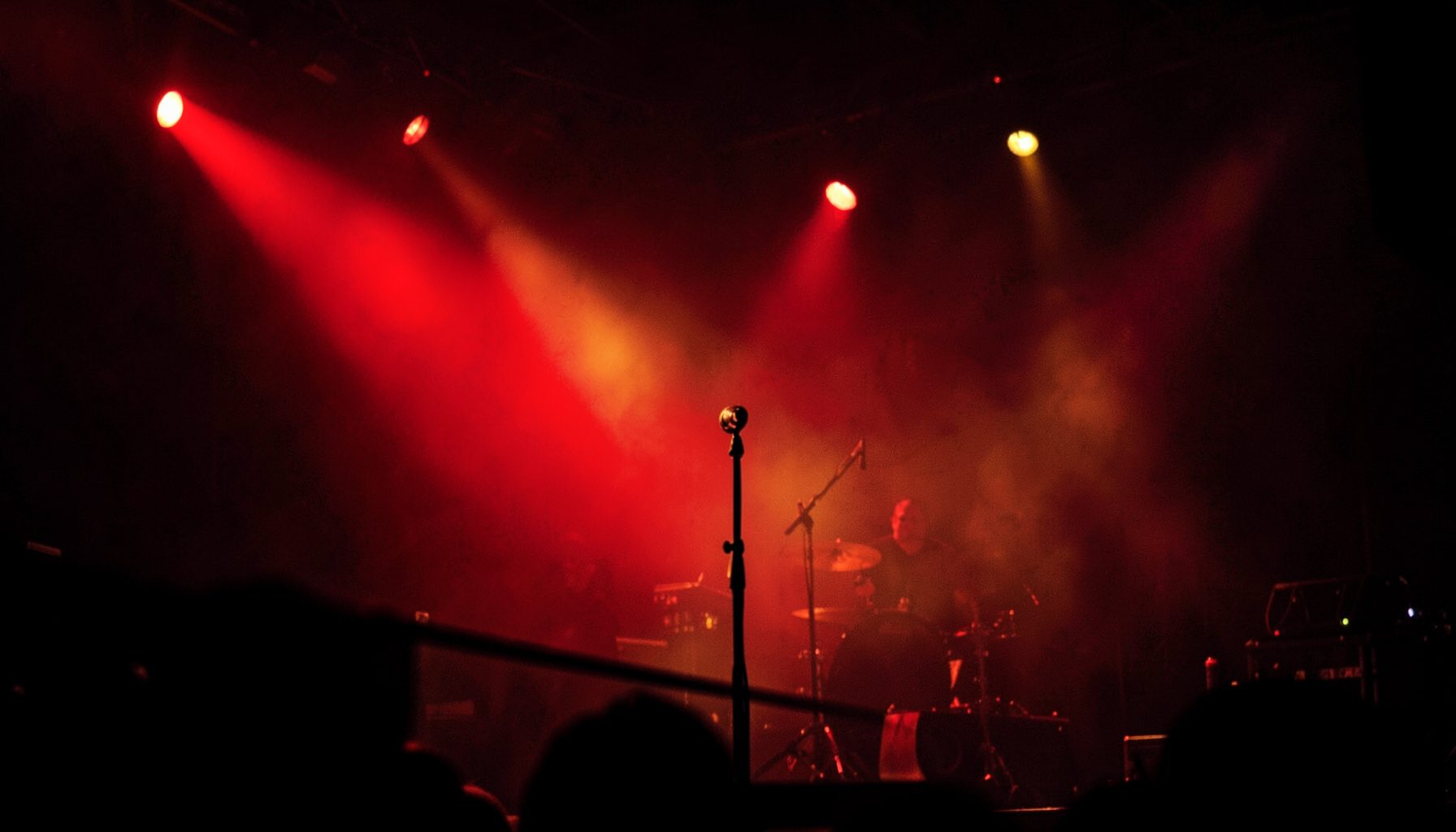 A dimly lit concert stage with red and amber spotlights cutting through haze, microphone stand silhouetted in the foreground, dark charcoal shadows wrapping the edges