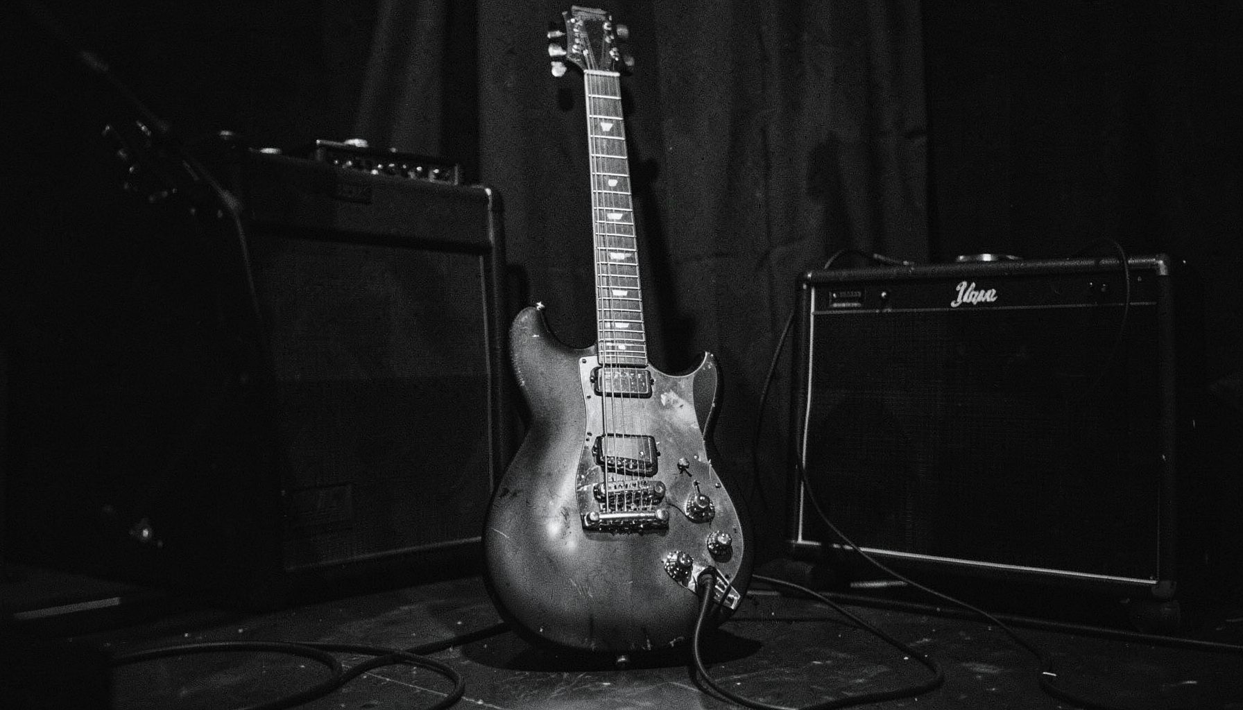 A moody black-and-white photograph of an electric guitar leaning against a stage amplifier, soft grain texture, dark charcoal shadows