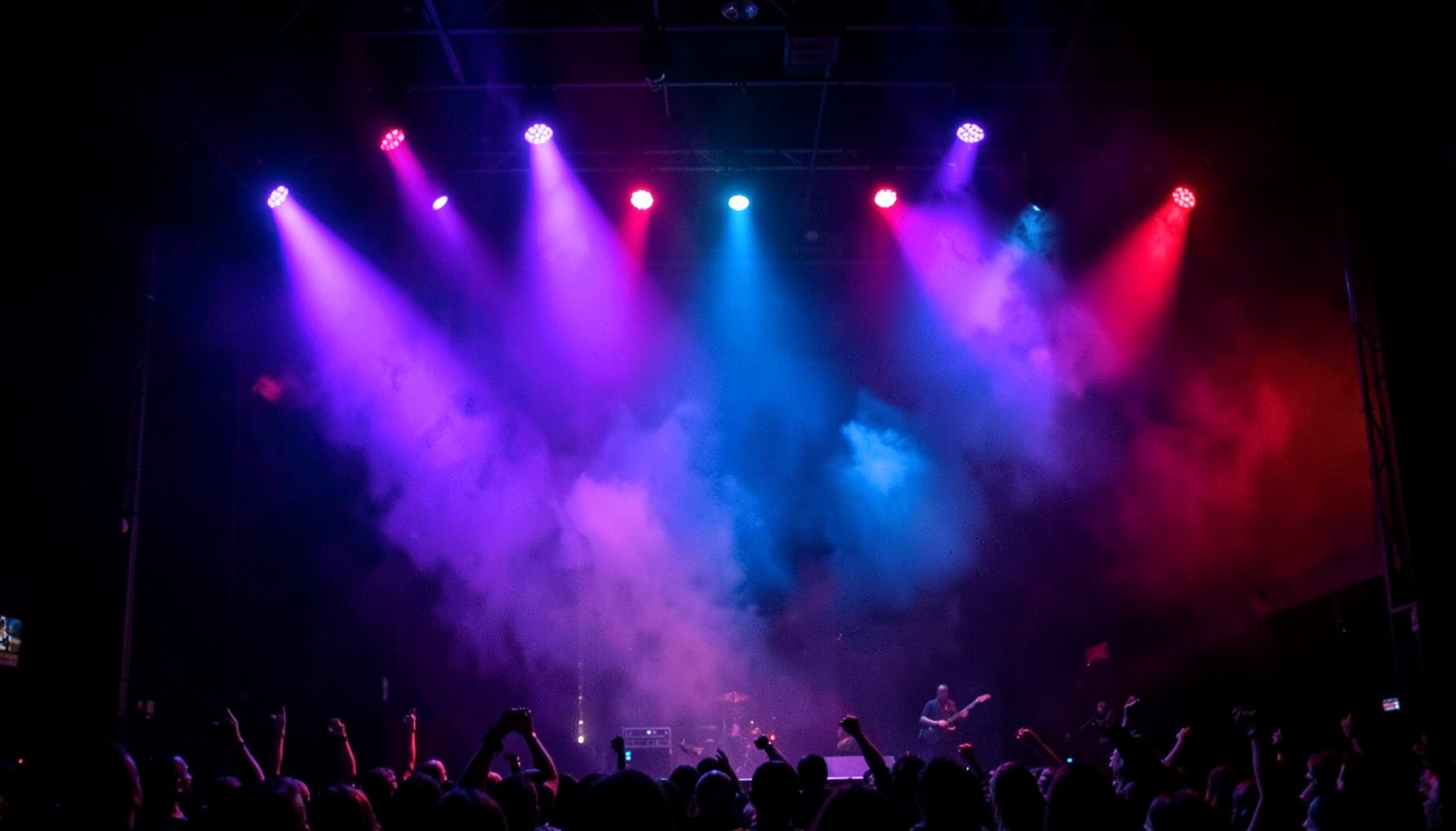 Dim stage with colorful concert spotlights in purple, blue, and red washing across a dark background, creating an energetic music atmosphere