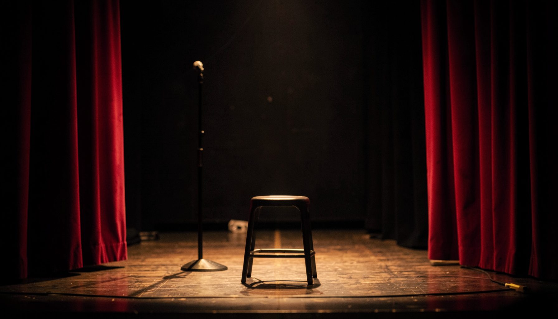 A warm, softly lit backstage scene with a microphone stand and an empty stool on a wooden stage, deep red curtains framing the sides, moody and intimate atmosphere